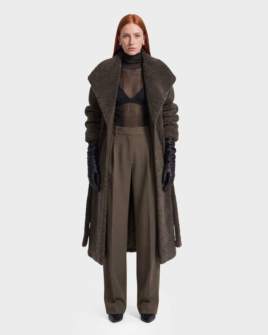 A person with long red hair wears a long brown textured coat, black gloves, a sheer black top with a visible bra, and the Rudsak FRANCIE - 8126873 Olive high-waisted wide-leg pants, standing against a plain white background. This striking look is available for pre-order now and is expected to ship by September 15th.