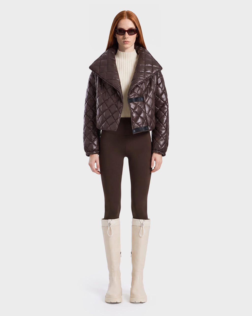 A woman with long red hair wears dark sunglasses, a shiny espresso Rudsak KALEE - 8124005 women's quilted light jacket, a cream turtleneck, brown pants, and tall white boots, standing against a plain white background.