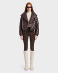 A woman with long red hair wears dark sunglasses, a shiny espresso Rudsak KALEE - 8124005 women's quilted light jacket, a cream turtleneck, brown pants, and tall white boots, standing against a plain white background.