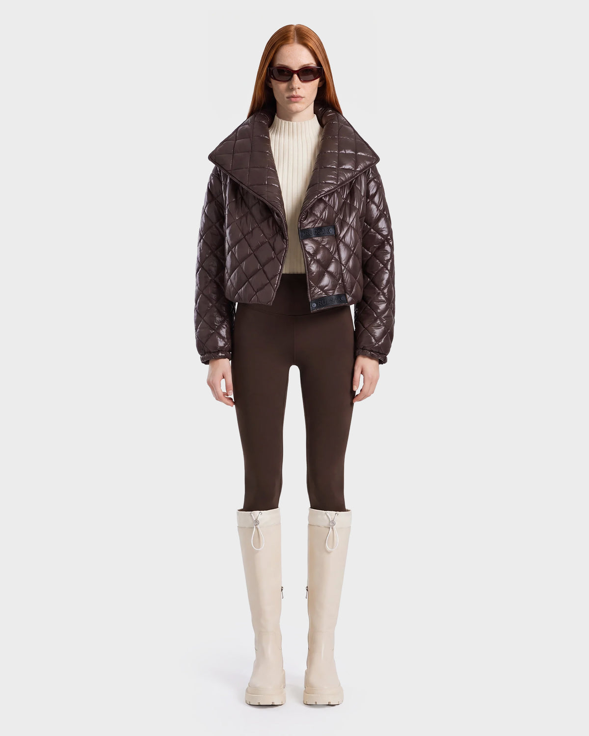A woman with long red hair wears dark sunglasses, a shiny espresso Rudsak KALEE - 8124005 women's quilted light jacket, a cream turtleneck, brown pants, and tall white boots, standing against a plain white background.