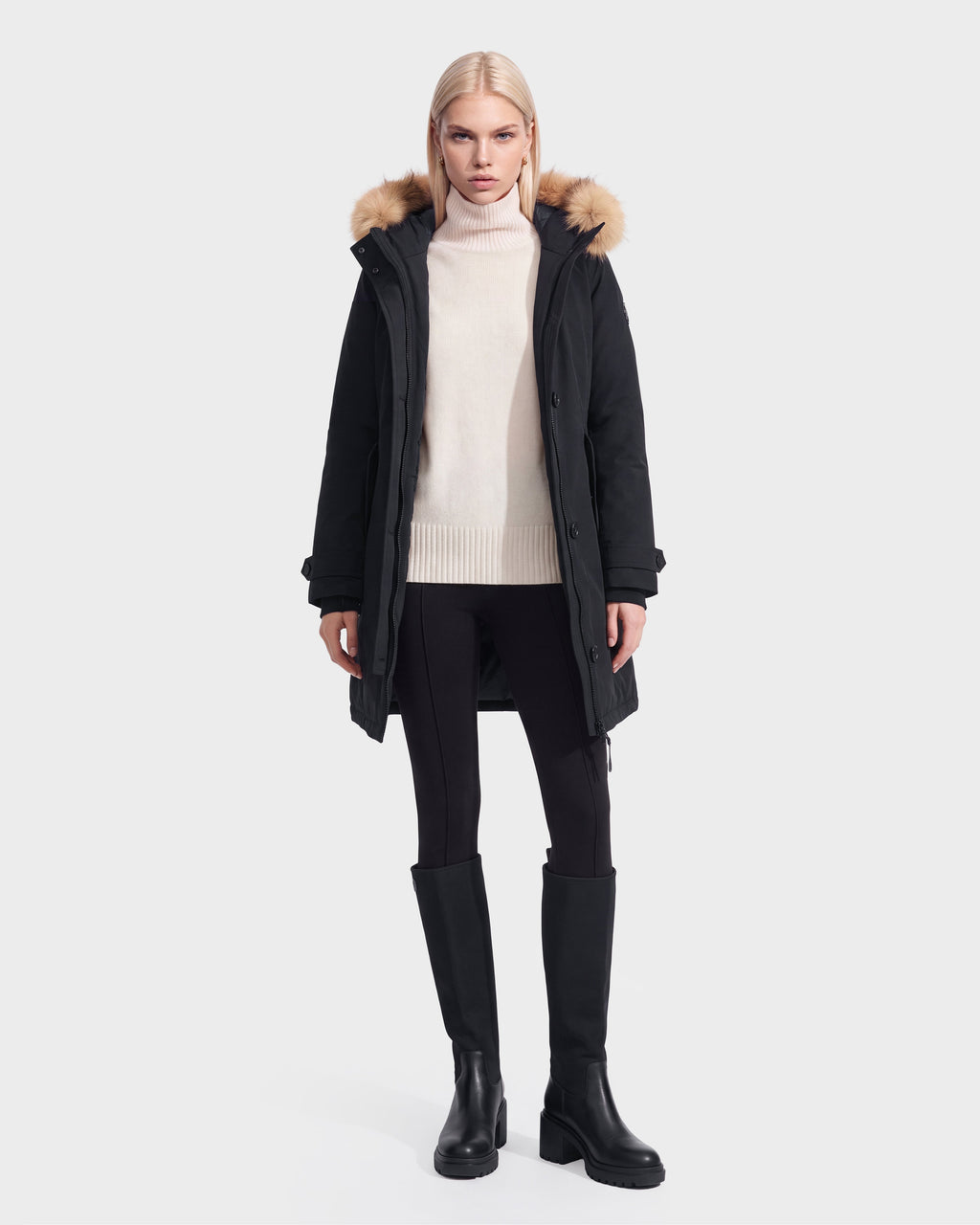 A woman models the Rudsak KEYSHIA - 8126517 Black down parka with a fur-trimmed hood, cream sweater, black leggings, and knee-high boots against a white background.