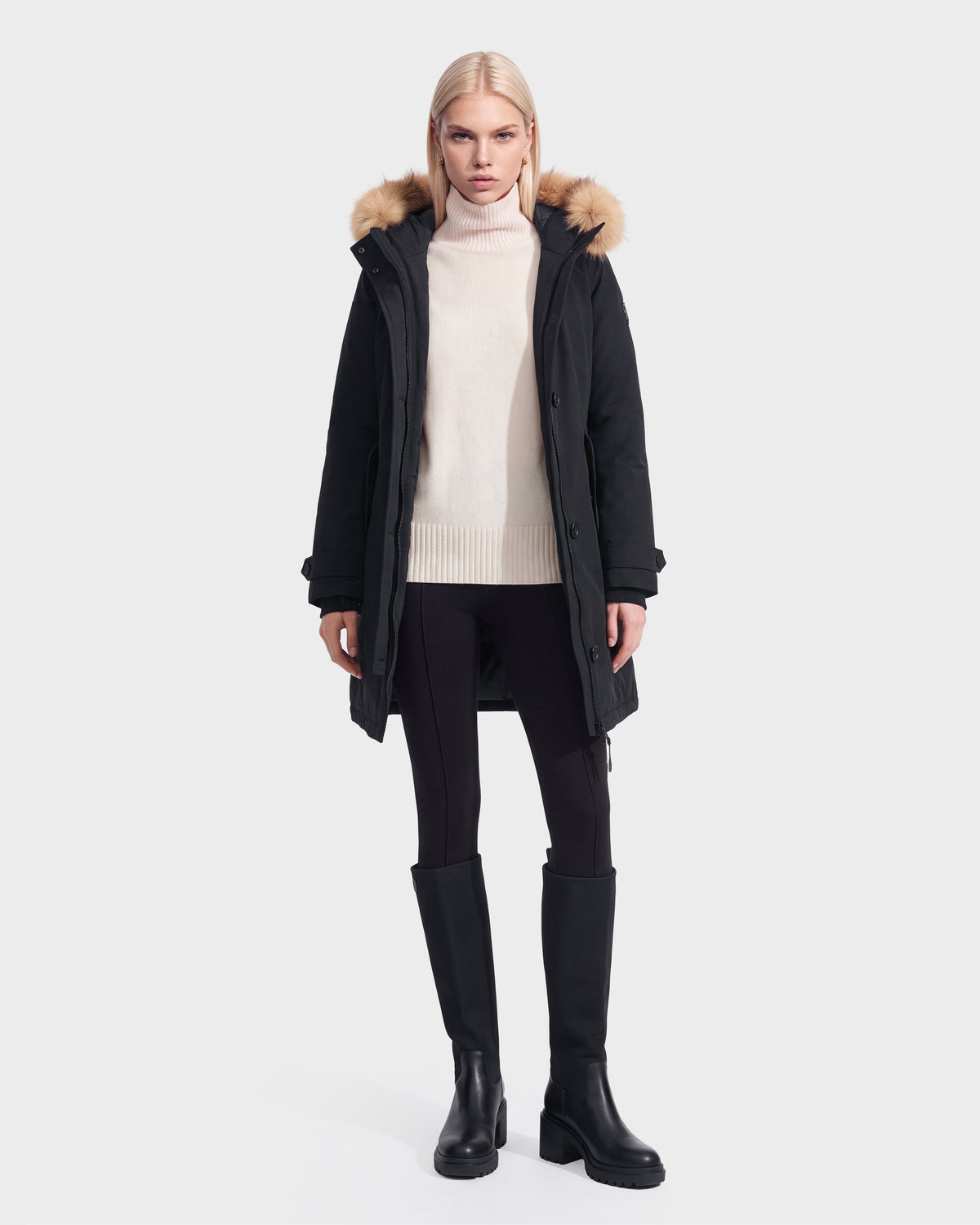 A woman models the Rudsak KEYSHIA - 8126517 Black down parka with a fur-trimmed hood, cream sweater, black leggings, and knee-high boots against a white background.