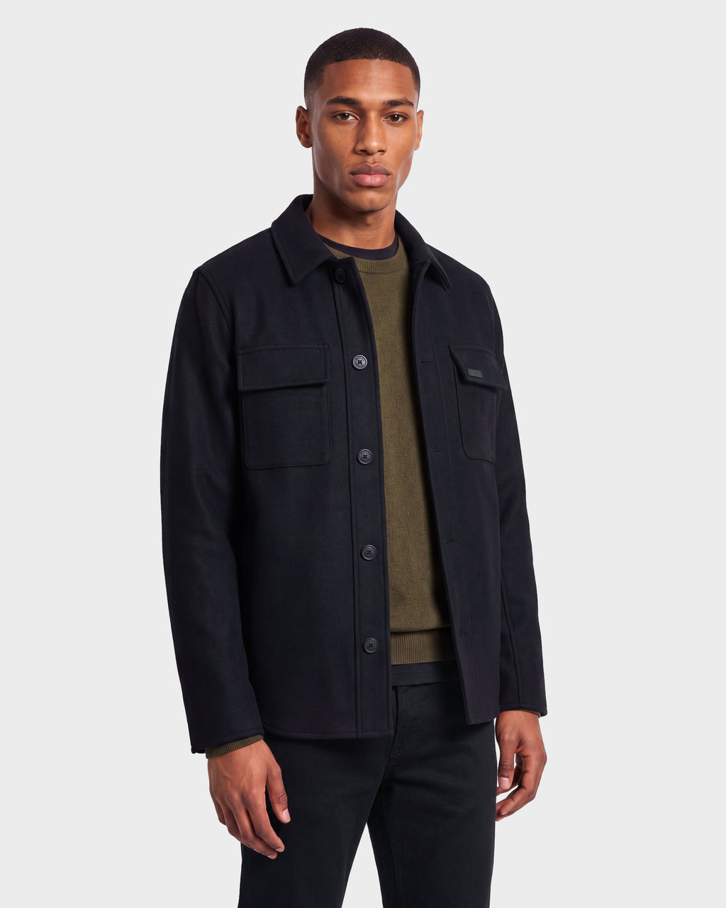A man stands against a plain background wearing the Rudsak MACKENZIE - 6126715 Black wool blend men’s shirt jacket over an olive green shirt and black pants, facing forward with a neutral expression—an ideal look for effortless transitional dressing.