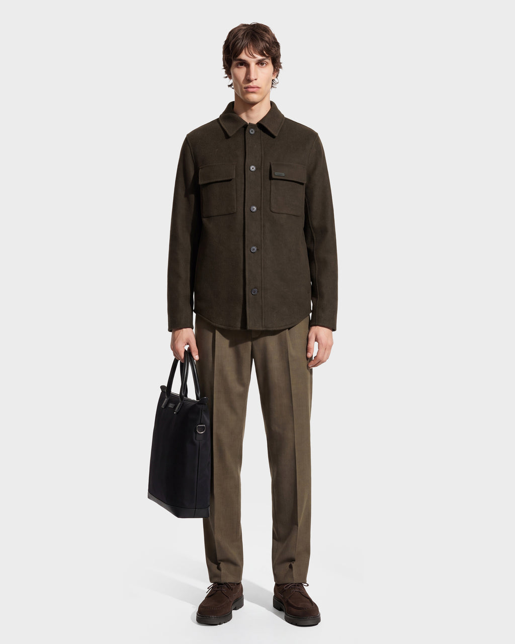 A man stands facing forward against a plain white background, wearing the Rudsak MACKENZIE - 6126715 Olive wool blend men’s shirt jacket with matching trousers and brown shoes, while holding a black tote bag in his left hand.