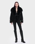 A person with straight, long brown hair wears the Rudsak MAEMI - 8126985 Black fur-collared coat paired with a black shirt, fitted black pants, and knee-high black boots, standing against a plain light gray background. This striking look is available for pre-order now and is expected to ship by September 15th.