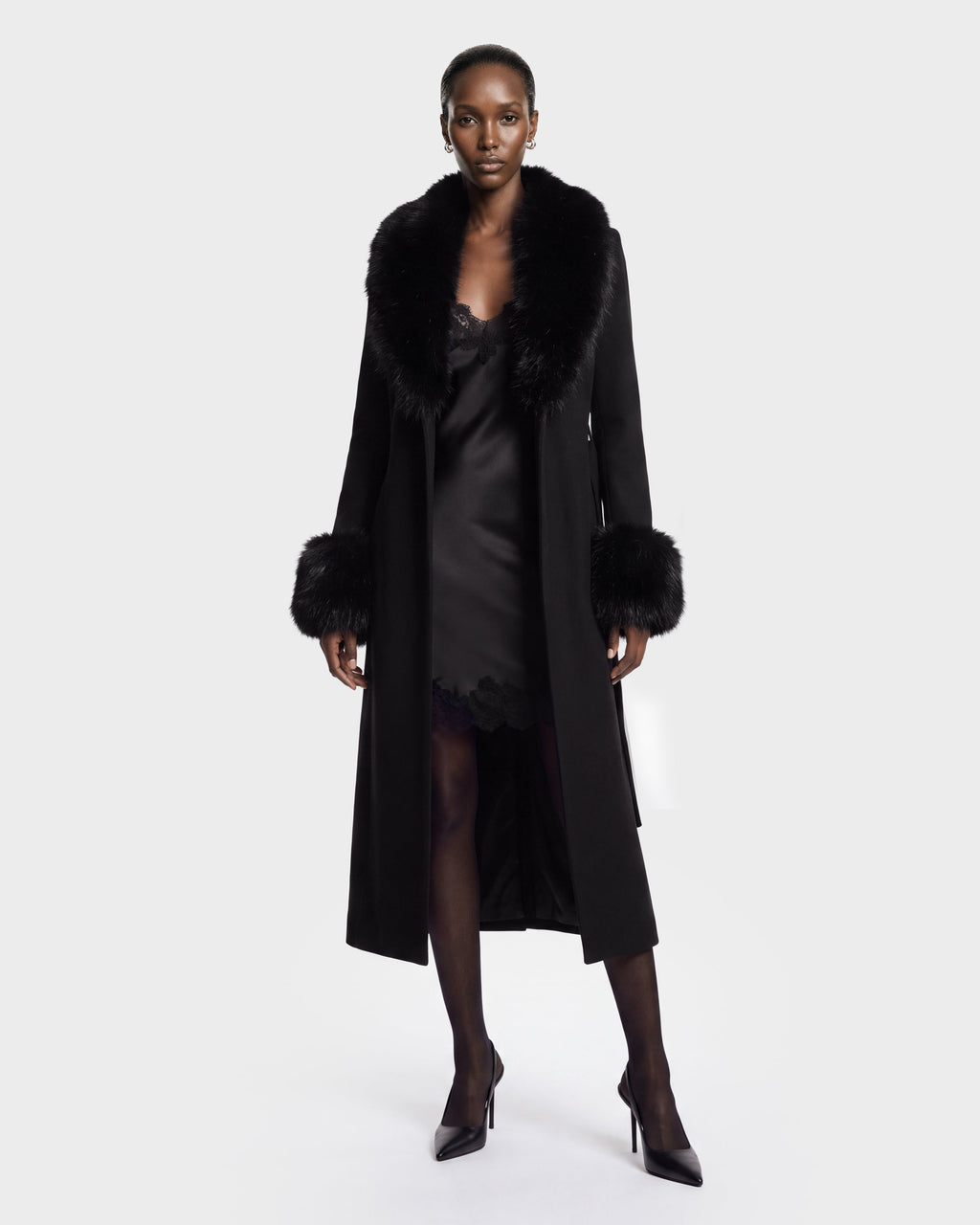 A woman stands against a plain background wearing the MARILLA WL - 8126737 Black coat by Rudsak, featuring luxurious fur trim at the collar and cuffs. She styles it with a black slip dress, sheer black tights, and black high heels. This stunning look is available for pre-order and is expected to ship by September 15th.