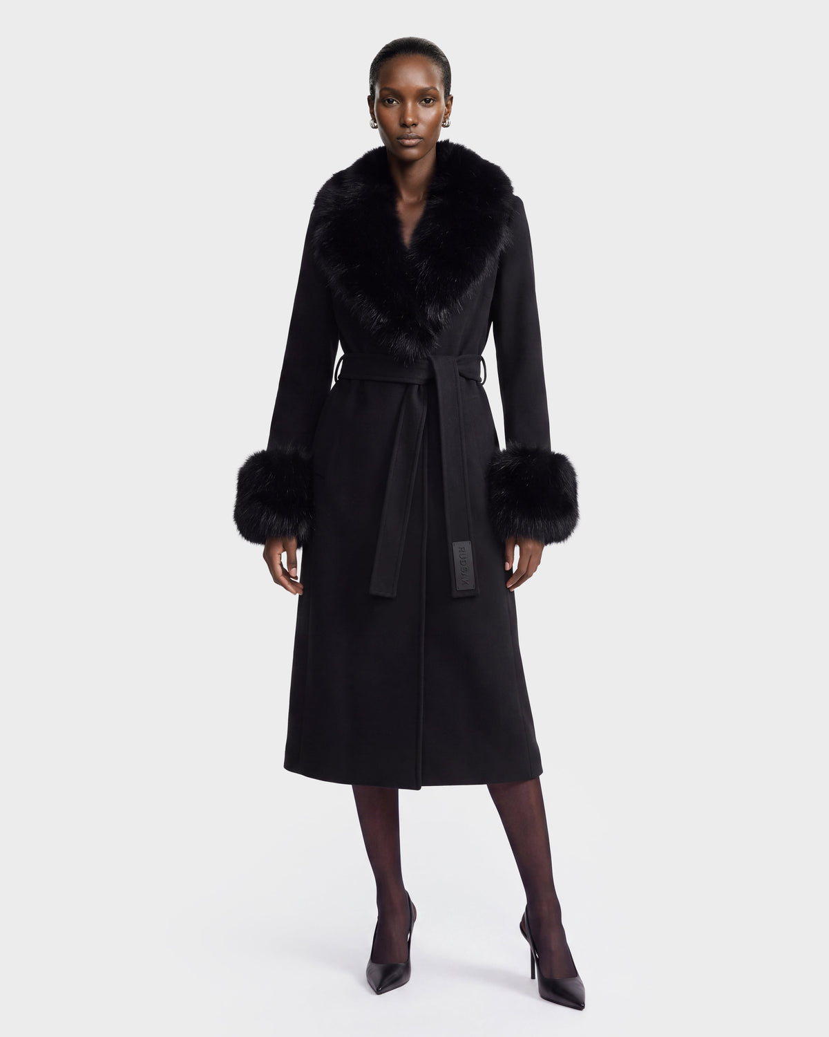 A person stands wearing the MARILLA WL - 8126737 Black coat by Rudsak, featuring a long silhouette with a fur collar and fur cuffs, belted at the waist and paired with black heels, against a plain light background. This elegant look is available for pre-order now and is expected to ship by September 15th.
