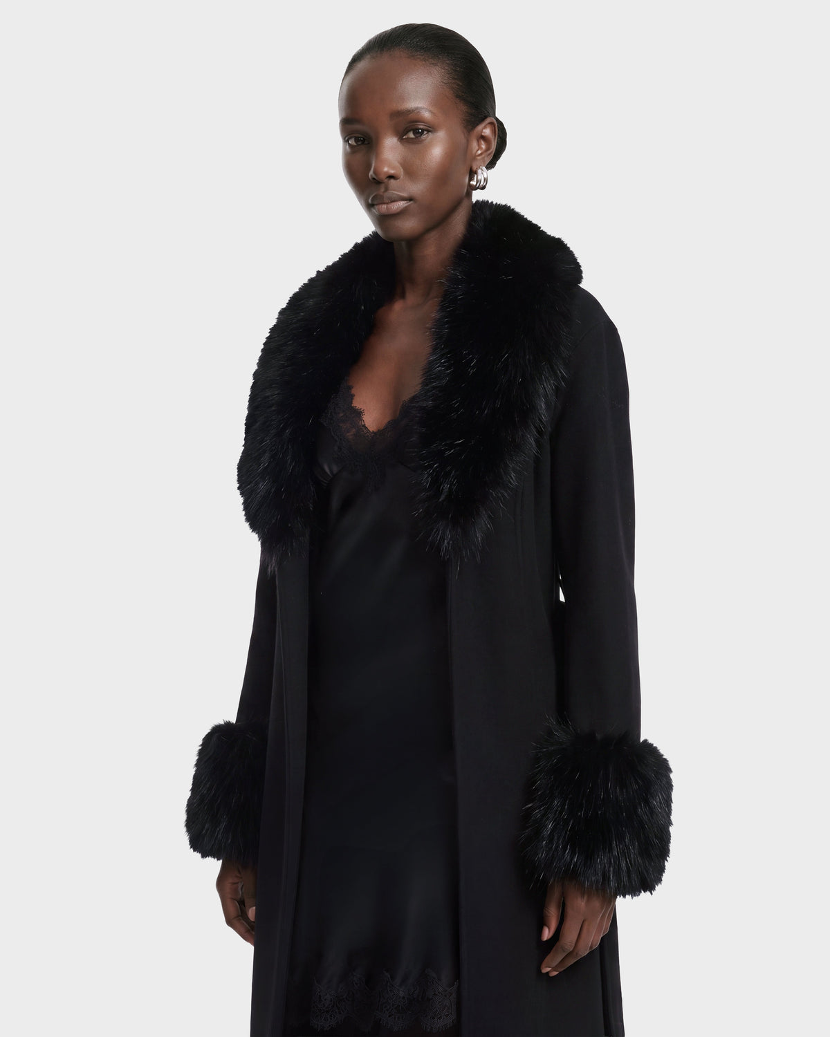 A person wearing the Rudsak MARILLA WL - 8126737 Black coat, featuring black fur trim on the collar and cuffs over a black dress, stands against a plain light background. This stylish look is available for pre-order and is expected to ship by September 15th.