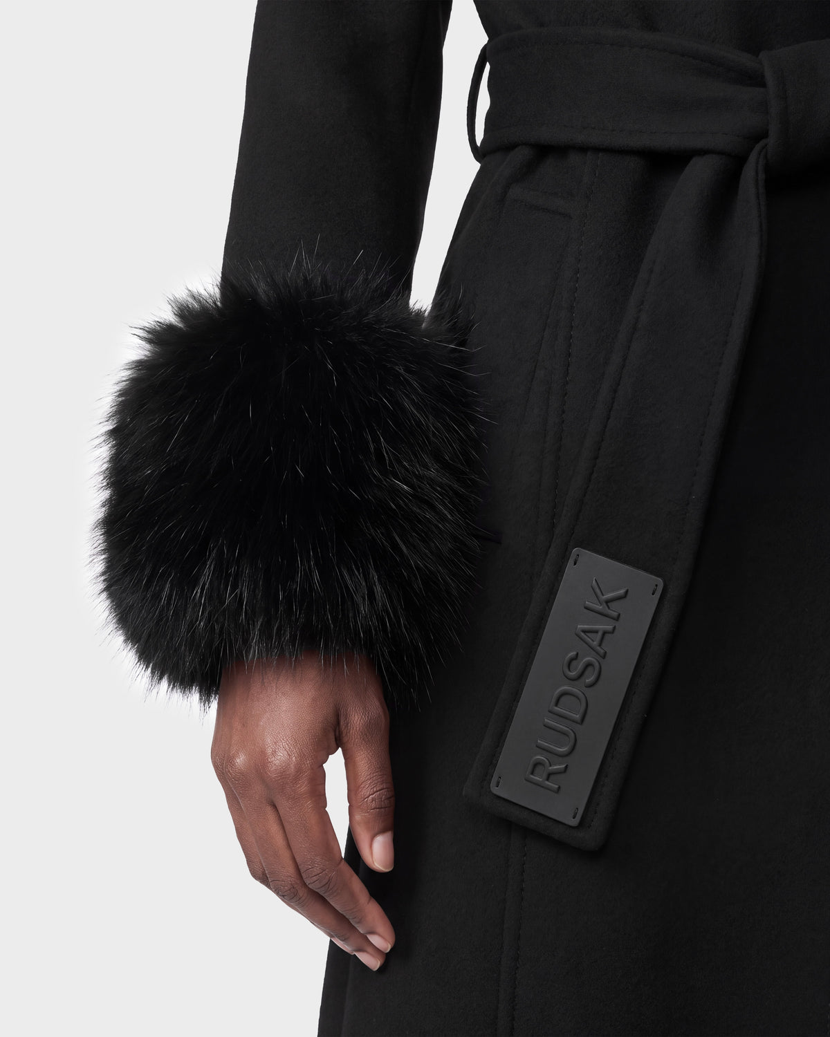 A person is wearing the MARILLA WL - 8126737 Black coat by Rudsak, featuring a fur cuff and belt tag. Their hand is visible by their side. This item is available for pre-order and is expected to ship by September 15th.