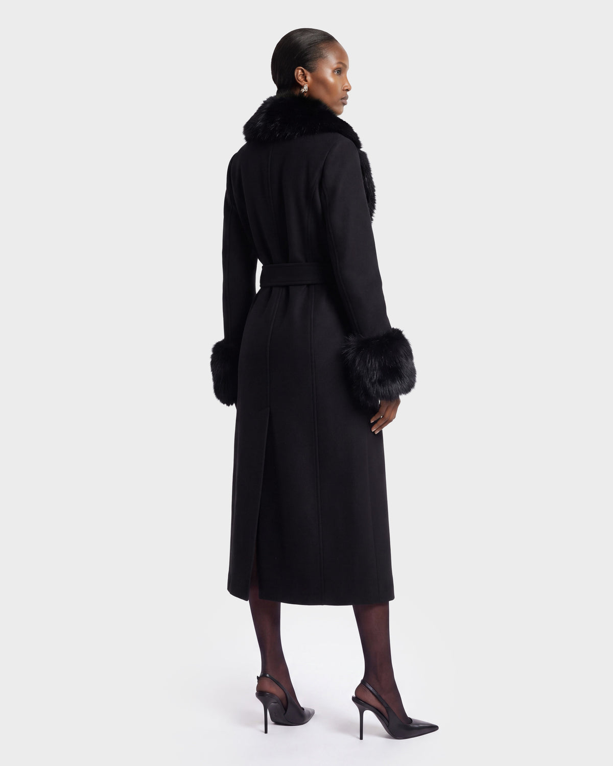 A woman stands facing away, wearing the MARILLA WL - 8126737 Black long coat by Rudsak, featuring a fur collar and cuffs, paired with black stockings and black high-heeled shoes against a plain white background. This item is available for pre-order and is expected to ship by September 15th.