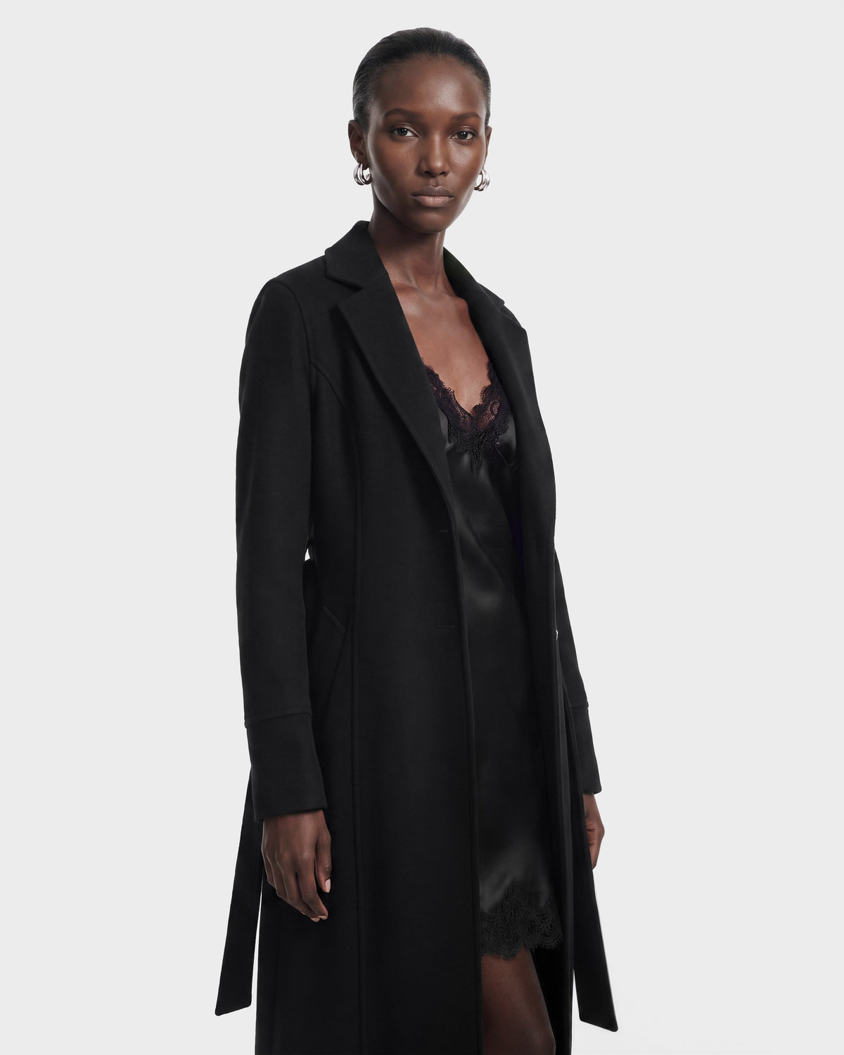 A woman stands against a plain background, wearing the Rudsak MARILLA WL - 8126737 Black, a long black coat layered over a black lace-trimmed dress. She has short hair, hoop earrings, and a calm expression. This elegant look is available for pre-order now and is expected to ship by September 15th.