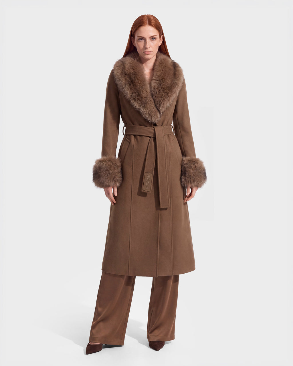 A woman stands facing forward wearing the Rudsak MARILLA WL - 8126737 Dark Caramel, a long brown coat with a wide fur collar and fur cuffs, tied at the waist. She has long straight red hair and is styled with brown wide-leg pants. Set against a plain white background, this striking piece is available for pre-order and is expected to ship by September 15th.