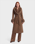 A woman stands facing forward wearing the Rudsak MARILLA WL - 8126737 Dark Caramel, a long brown coat with a wide fur collar and fur cuffs, tied at the waist. She has long straight red hair and is styled with brown wide-leg pants. Set against a plain white background, this striking piece is available for pre-order and is expected to ship by September 15th.