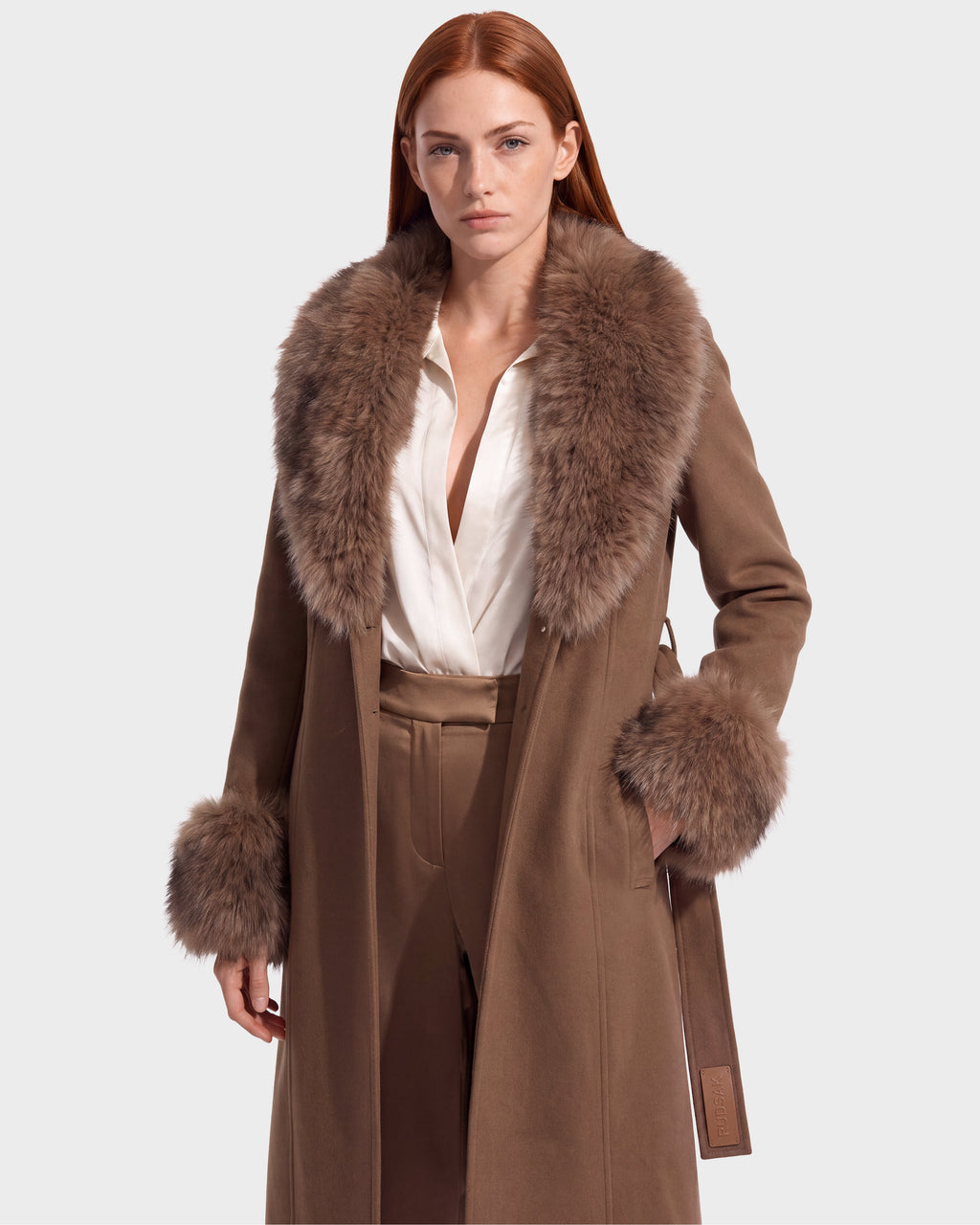 A woman with long red hair models the Rudsak MARILLA WL - 8126737 Dark Caramel coat, featuring a large, fluffy brown fur collar and cuffs, worn over a white blouse and matching tan pants against a plain background. This striking look is available for pre-order now and is expected to ship by September 15th.