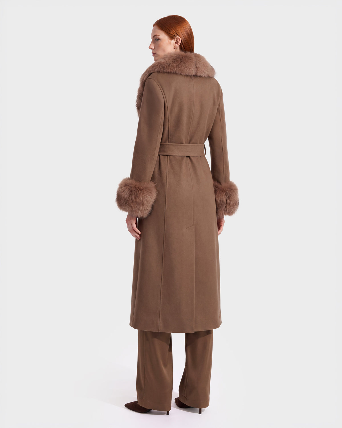 A woman with red hair wears the Rudsak MARILLA WL - 8126737 Dark Caramel, a long brown coat with fur-trimmed collar and cuffs, shown from behind. She pairs it with matching brown pants and dark shoes against a plain white background. This stylish look is available for pre-order now and is expected to ship by September 15th.