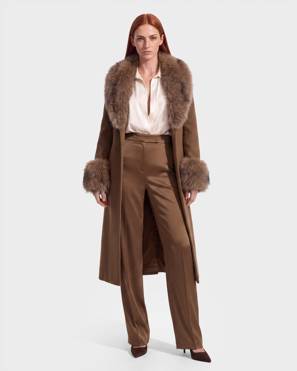 Woman with long red hair wearing the Rudsak MARILLA WL - 8126737 Dark Caramel coat, featuring a fur collar and cuffs, paired with a white blouse, tan high-waisted trousers, and brown pointed shoes, standing against a plain light background. Now available for pre-order and expected to ship by September 15th.