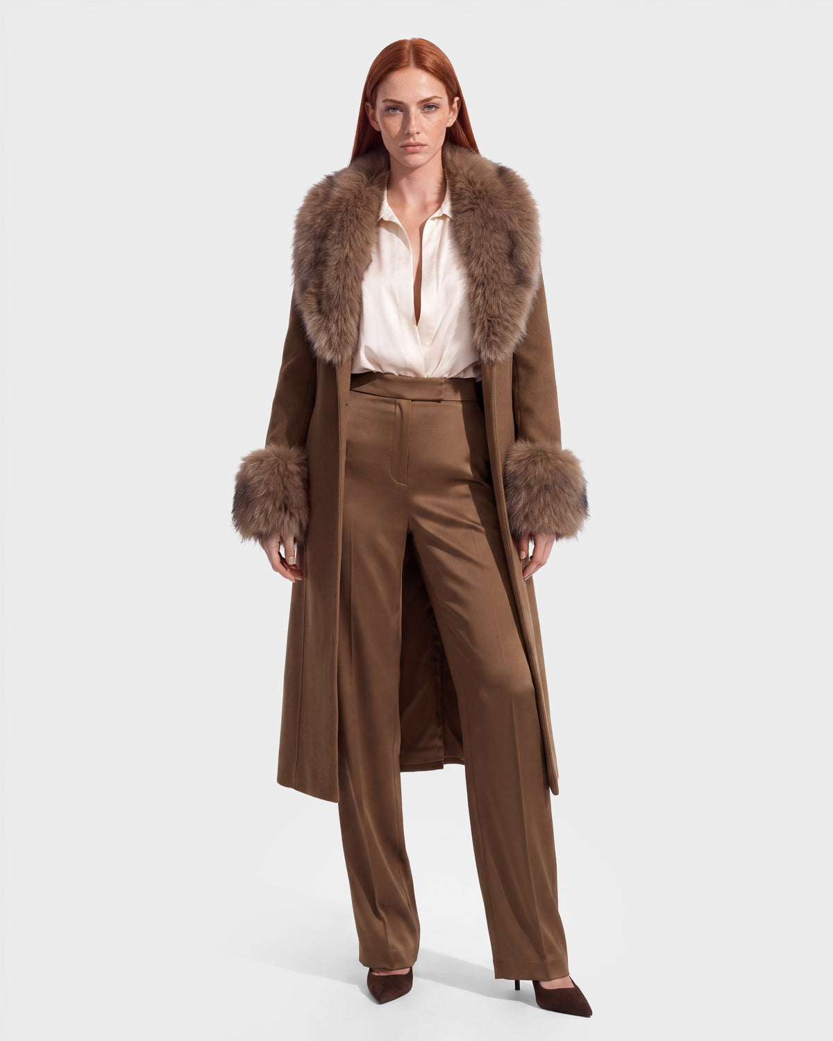 Woman with long red hair wearing the Rudsak MARILLA WL - 8126737 Dark Caramel coat, featuring a fur collar and cuffs, paired with a white blouse, tan high-waisted trousers, and brown pointed shoes, standing against a plain light background. Now available for pre-order and expected to ship by September 15th.