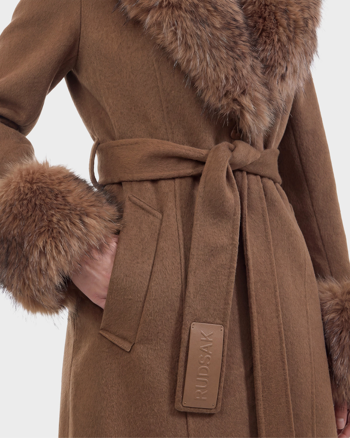 A person wearing the Rudsak MARILLA WL - 8126737 coat in Dark Caramel, featuring fur trim on the collar and cuffs and a belted waist with a visible brand tag. This stylish piece is available for pre-order now and is expected to ship by September 15th.