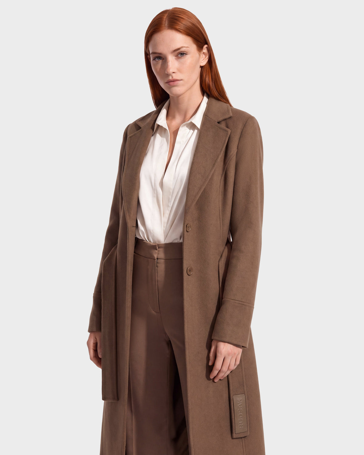 A woman with long red hair wears the Rudsak MARILLA WL - 8126737 coat in Dark Caramel over a white blouse and brown pants, standing against a plain light background. This stylish look is now available for pre-order and is expected to ship by September 15th.