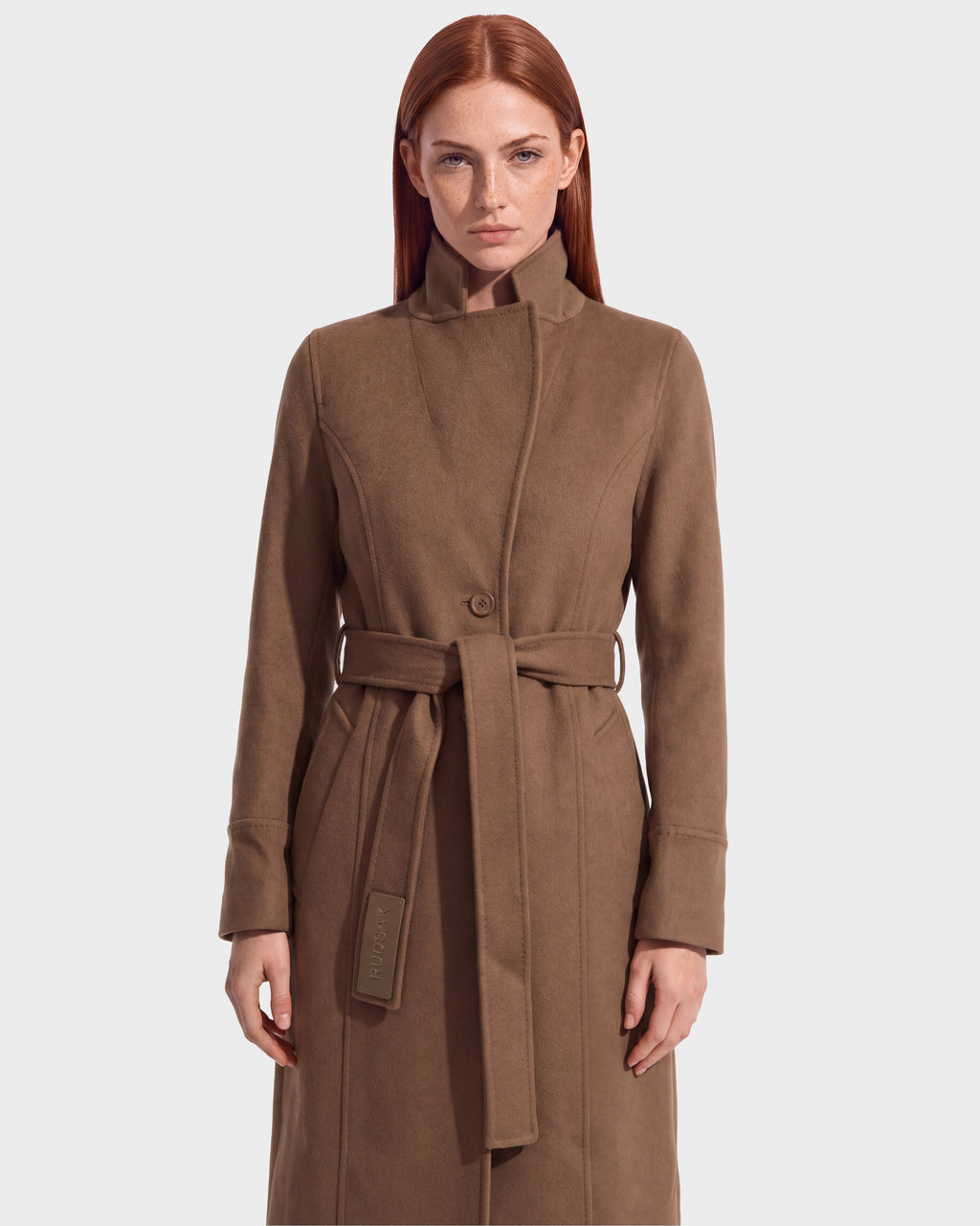 A woman with long red hair wears the Rudsak MARILLA WL - 8126737 Dark Caramel brown belted coat, standing against a plain light gray background and facing forward with a neutral expression. Available for pre-order now; expected to ship by September 15th.