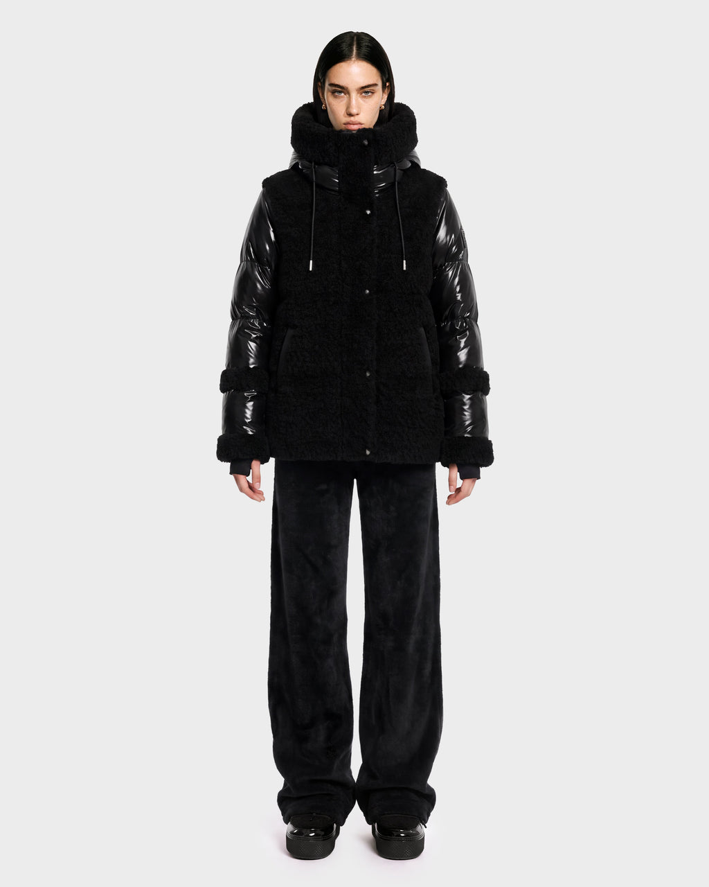 A person stands facing forward against a plain light gray background, wearing the Rudsak MARISSA - 8126728 Black down jacket featuring shiny sleeves and a fuzzy, winter-inspired texture, paired with loose black pants and black shoes.