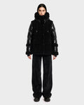 A person stands facing forward against a plain light gray background, wearing the Rudsak MARISSA - 8126728 Black down jacket featuring shiny sleeves and a fuzzy, winter-inspired texture, paired with loose black pants and black shoes.