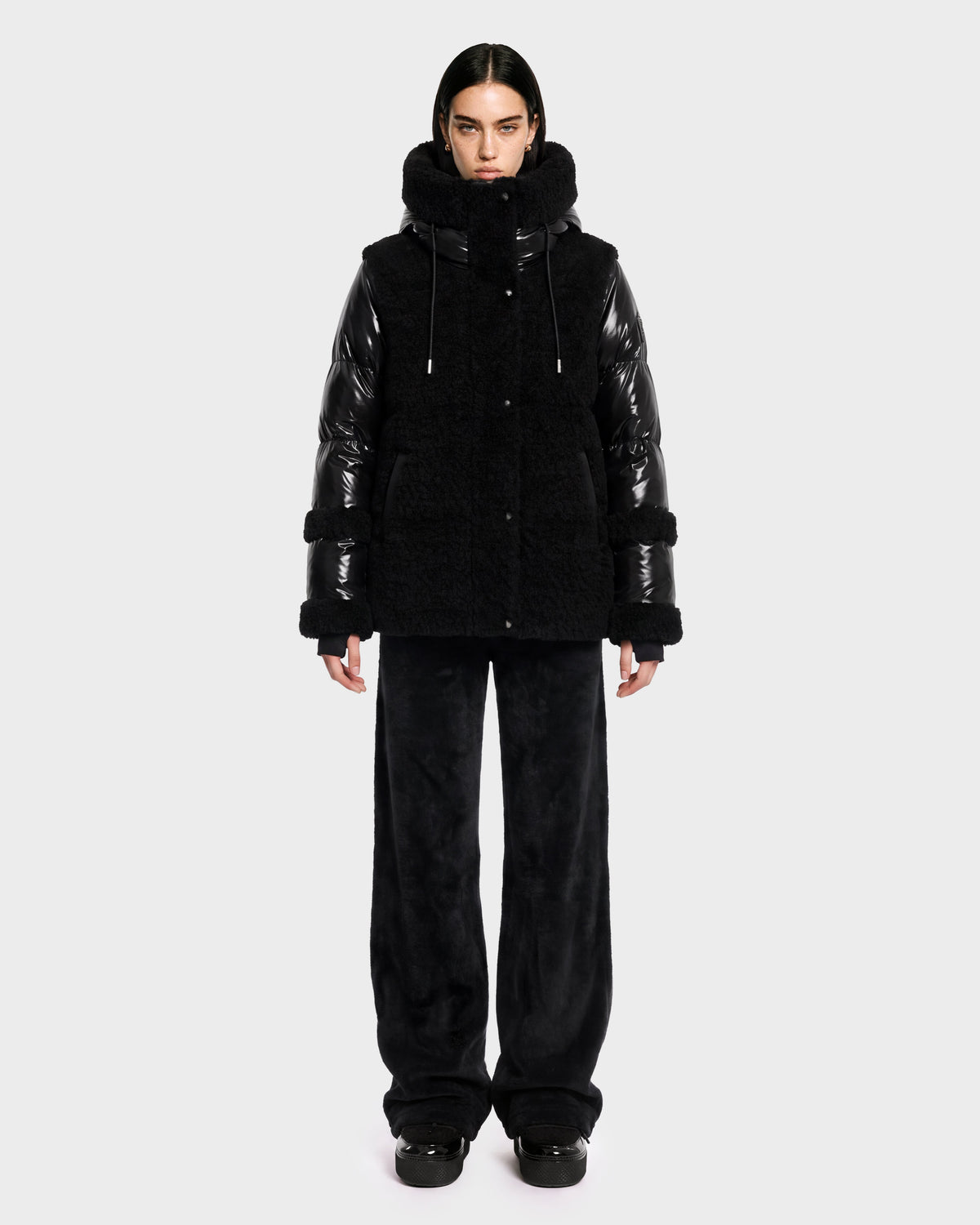 A person stands facing forward against a plain light gray background, wearing the Rudsak MARISSA - 8126728 Black down jacket featuring shiny sleeves and a fuzzy, winter-inspired texture, paired with loose black pants and black shoes.