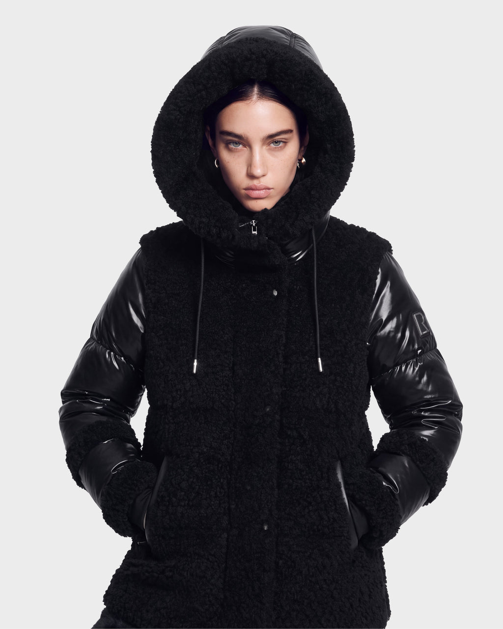 A person wearing the MARISSA - 8126728 Black winter coat by Rudsak, featuring a hood with faux fur trim and sleek, shiny material, stylishly poses with hands in pockets against a plain light background while looking directly at the camera.