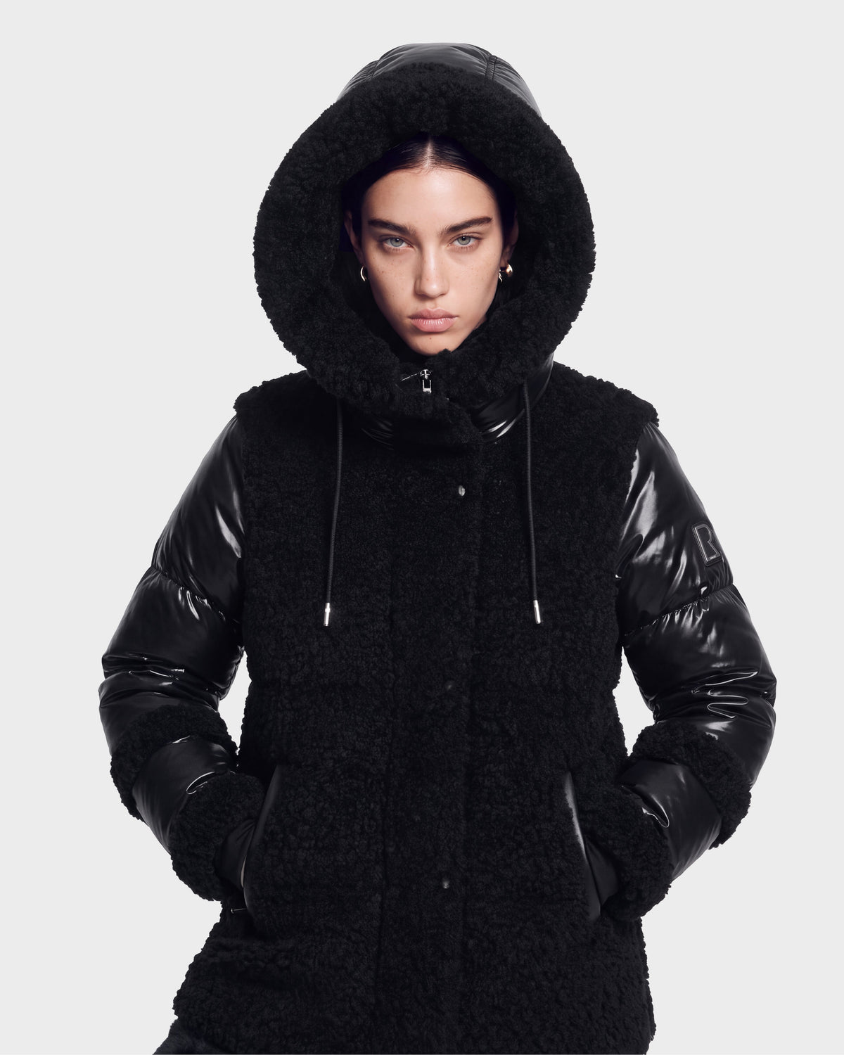 A person wearing the MARISSA - 8126728 Black winter coat by Rudsak, featuring a hood with faux fur trim and sleek, shiny material, stylishly poses with hands in pockets against a plain light background while looking directly at the camera.