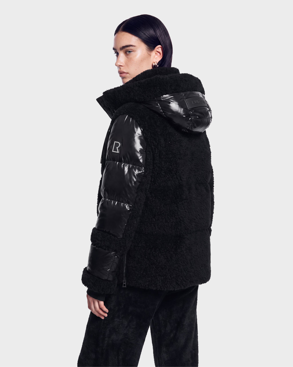 A person with dark hair is shown from the back, standing against a plain light background and looking over their shoulder while wearing the Rudsak MARISSA - 8126728 Black, a textured and shiny hooded down jacket.