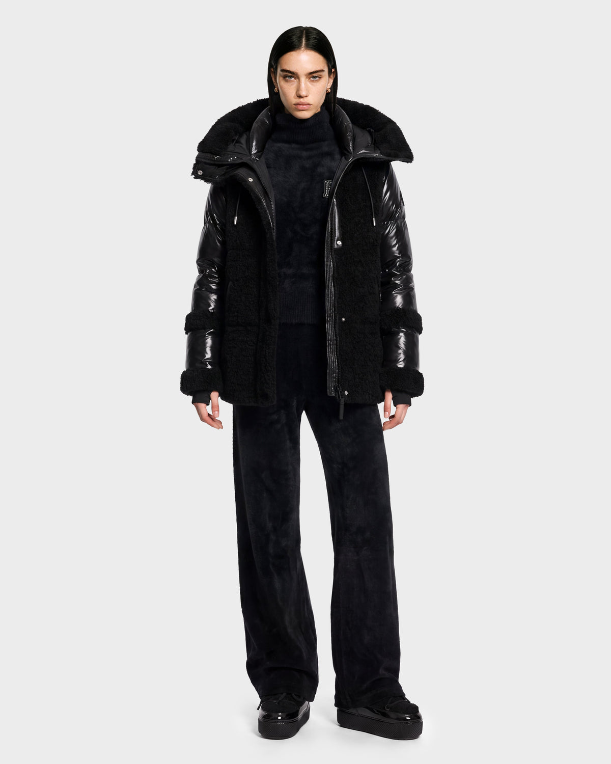 A person stands facing forward against a plain light gray background, dressed in stylish winter outerwear from Rudsak—the MARISSA - 8126728 Black puffer down jacket featuring faux fur details, paired with a black turtleneck, wide-leg black pants, and black shoes.