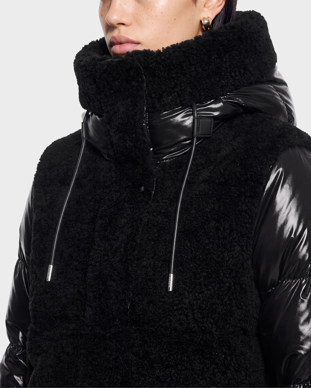 A person wearing the Rudsak MARISSA - 8126728 Black coat, featuring a high textured collar, shiny sleeves, and drawstring details, looks slightly off to the side with only the lower half of their face visible.