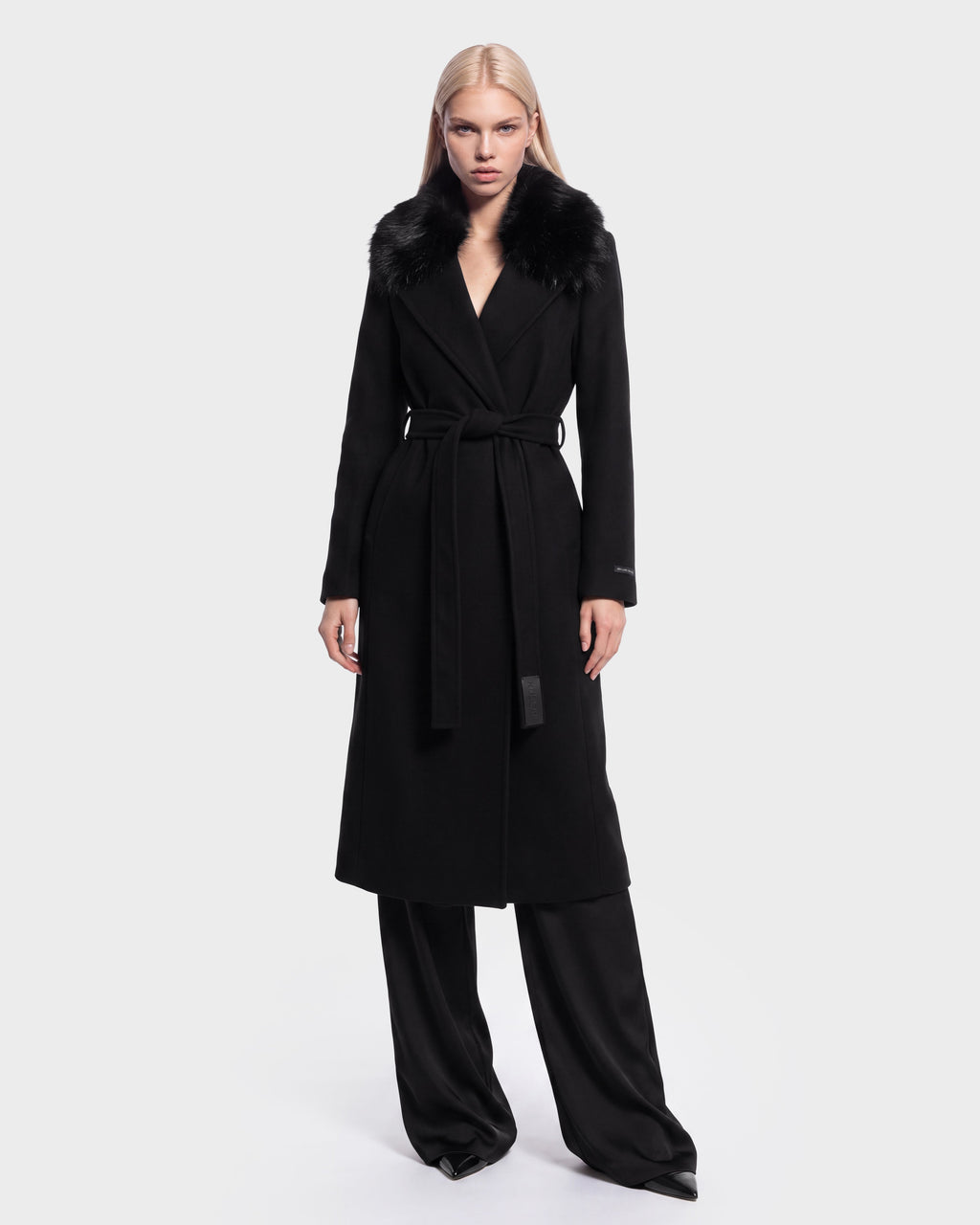 A woman wears the Rudsak MEL - 8126525 Black long coat, featuring a fur collar and waist tie, layered over black wide-leg pants and standing against a plain white background. This stylish coat is available for pre-order and is expected to ship by September 15th.