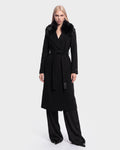 A woman wears the Rudsak MEL - 8126525 Black long coat, featuring a fur collar and waist tie, layered over black wide-leg pants and standing against a plain white background. This stylish coat is available for pre-order and is expected to ship by September 15th.