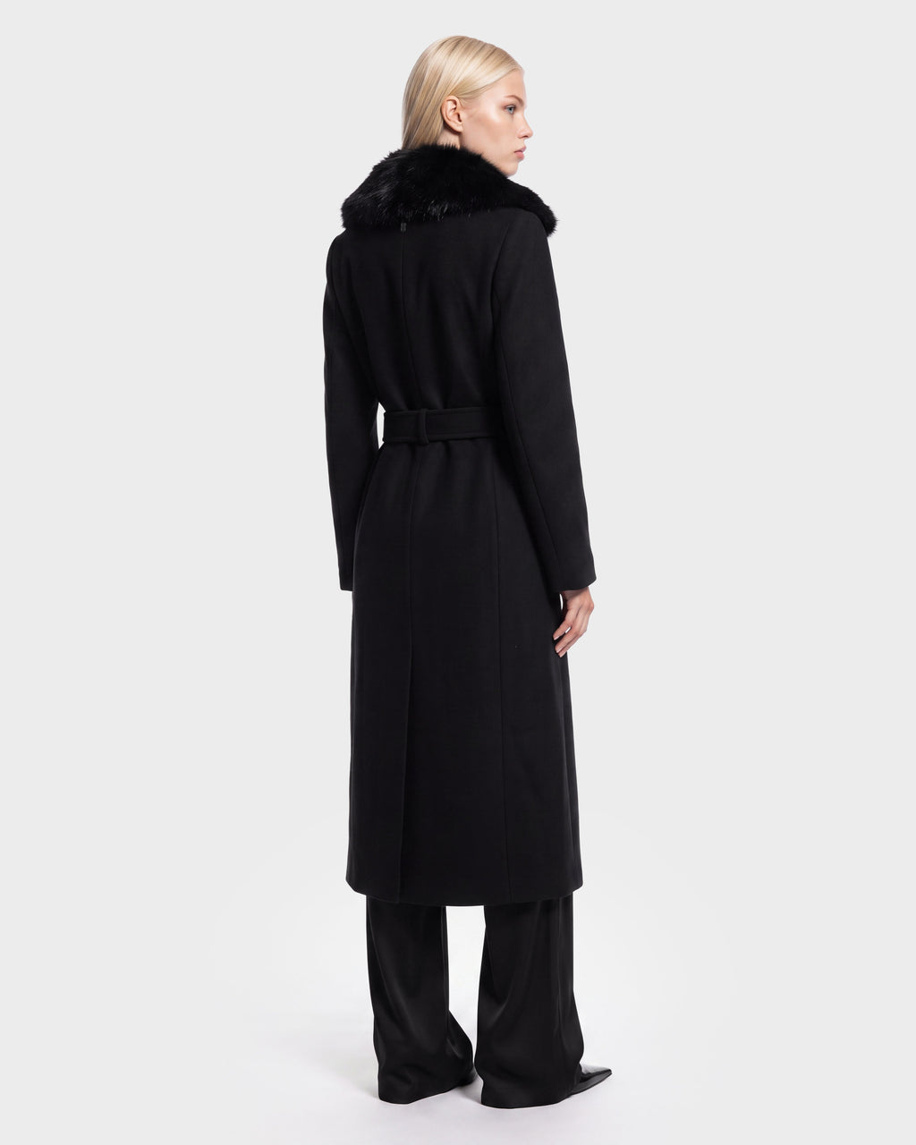 A woman with long blonde hair stands facing away against a plain white background, wearing the Rudsak MEL - 8126525 Black long coat featuring a faux fur collar, paired with black pants and black shoes. This stylish look is available for pre-order and is expected to ship by September 15th.
