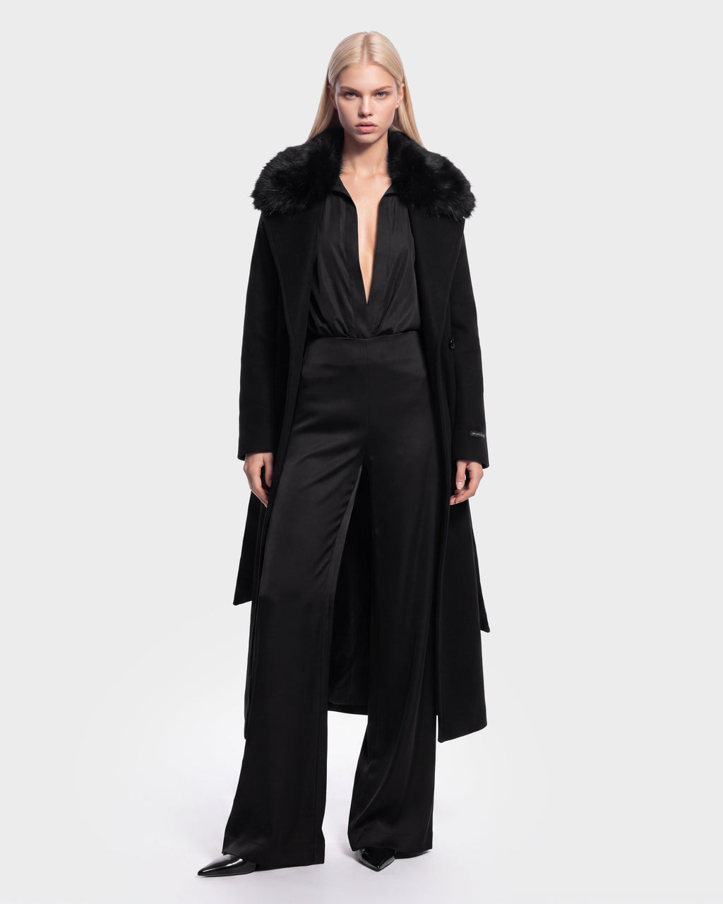 A woman with long blonde hair wears the Rudsak MEL - 8126525 Black coat with a fur collar over a deep V-neck black jumpsuit and pointed black shoes, standing against a plain white background. This stylish look is available for pre-order and is expected to ship by September 15th.
