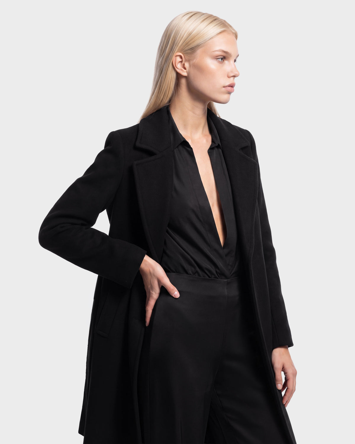 Model wears the MEL - 8126525 Black coat by Rudsak over a deep V-neck black blouse and black pants, posing against a plain light background with one hand on her hip. This stylish look is now available for pre-order from Rudsak and is expected to ship by September 15th.