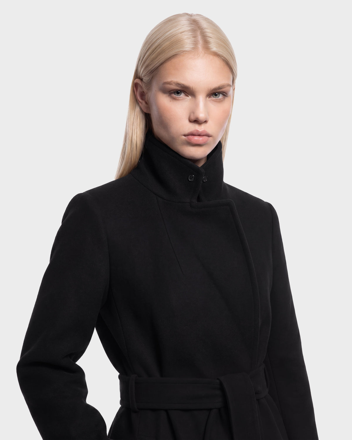 A person with long, straight blonde hair wears the Rudsak MEL - 8126525 Black belted coat with a high collar, looking directly at the camera against a plain light background. This striking Rudsak piece is available for pre-order and is expected to ship by September 15th.