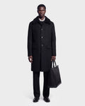 A man stands against a plain light gray background, looking straight at the camera, wearing the Rudsak NICO - 6126717 Black men’s long wool coat with a removable faux-shearling collar, paired with black pants, black shoes, and holding a black tote bag.