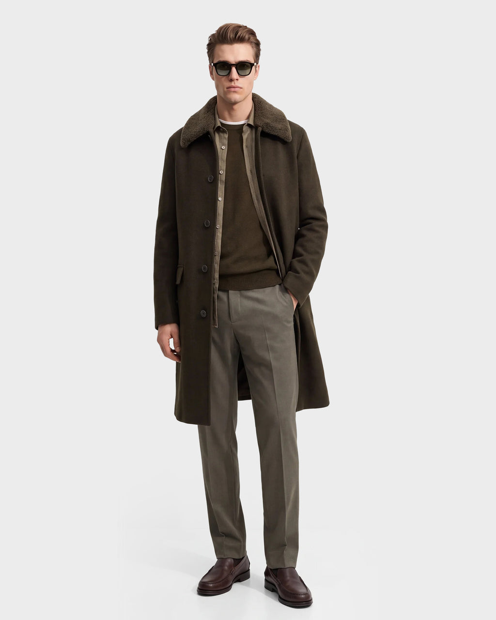 A man wearing sunglasses, Rudsak’s NICO - 6126717 Olive wool blend coat with a shearling collar, an olive sweater, khaki trousers, and brown shoes stands with one hand in his pocket against a plain light background.