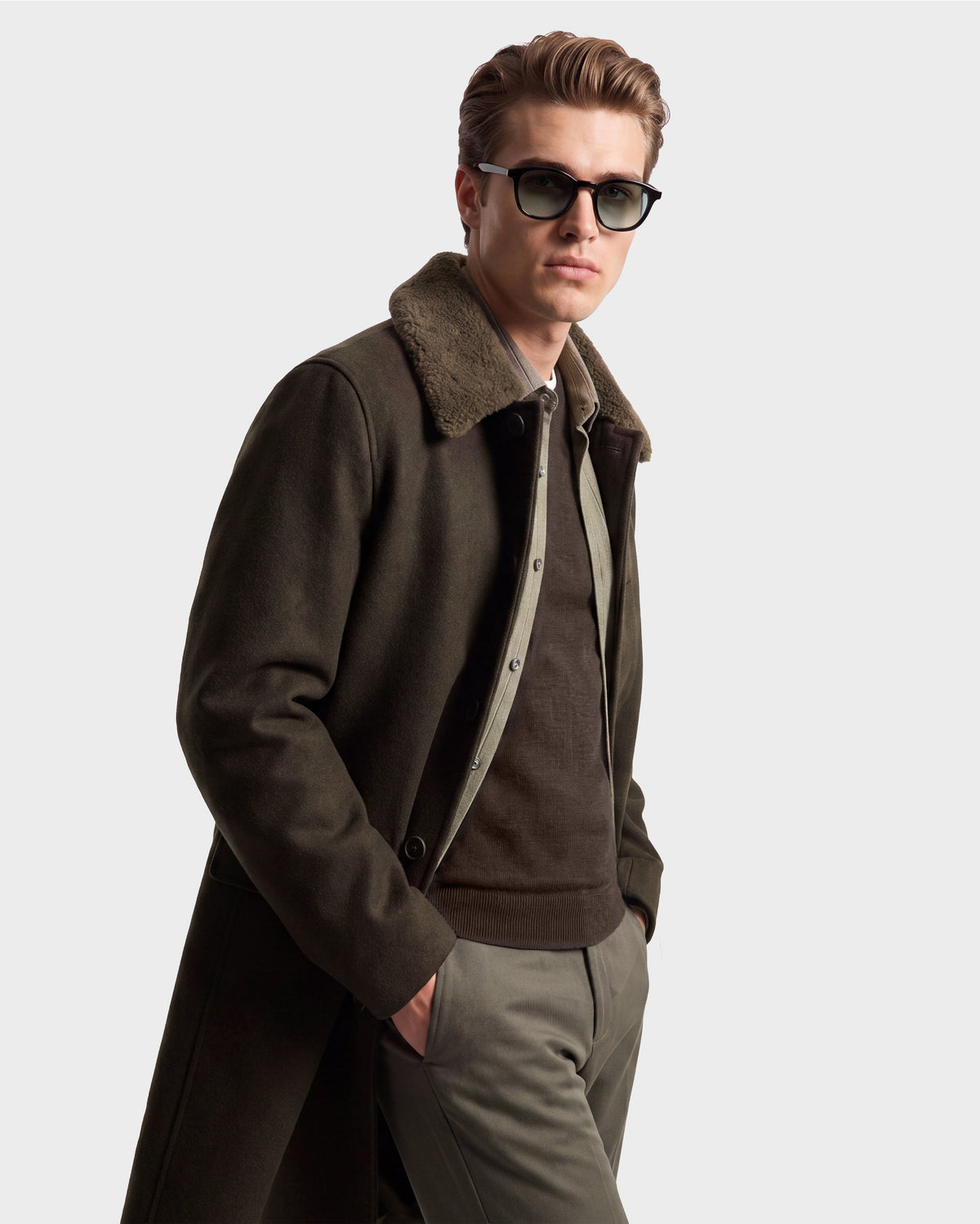 A man wearing sunglasses, an olive green NICO - 6126717 jacket by Rudsak with a fur collar, a brown sweater, and green pants stands with one hand in his pocket against a plain light background.