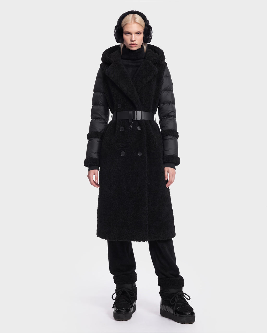 A woman is wearing the Rudsak NOELLE - 8126971 Black double-breasted coat, featuring textured fabric and subtle faux fur accents. She pairs it with black pants, black boots, and earmuffs, standing against a plain white background with a neutral expression.