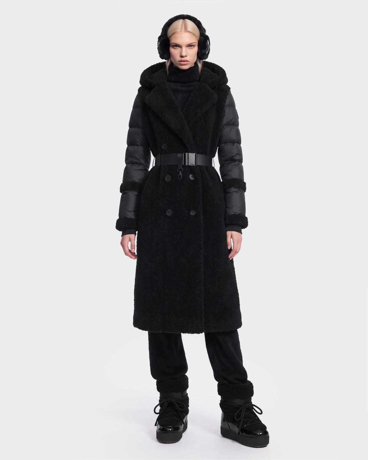A woman is wearing the Rudsak NOELLE - 8126971 Black double-breasted coat, featuring textured fabric and subtle faux fur accents. She pairs it with black pants, black boots, and earmuffs, standing against a plain white background with a neutral expression.