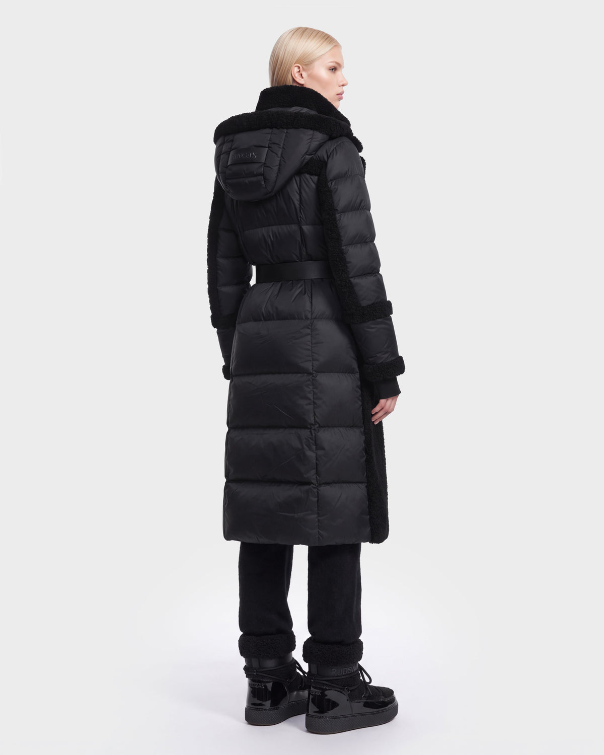 A person with straight blond hair is seen from behind against a plain white background, wearing the Rudsak NOELLE - 8126971 Black long hooded puffer coat with a belt and down insulation, paired with black pants and black shoes.