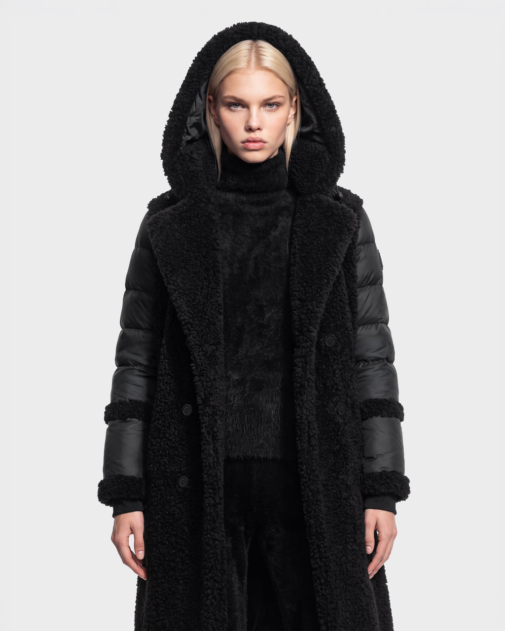 A woman with straight blond hair is shown standing against a plain white background, facing forward with a neutral expression, wearing the NOELLE - 8126971 Black long hooded coat by Rudsak. The coat features down insulation, a faux fur texture, and leather sleeves.