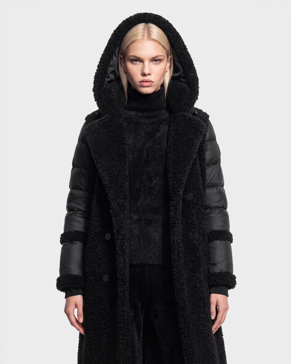 A woman with straight blond hair is shown standing against a plain white background, facing forward with a neutral expression, wearing the NOELLE - 8126971 Black long hooded coat by Rudsak. The coat features down insulation, a faux fur texture, and leather sleeves.