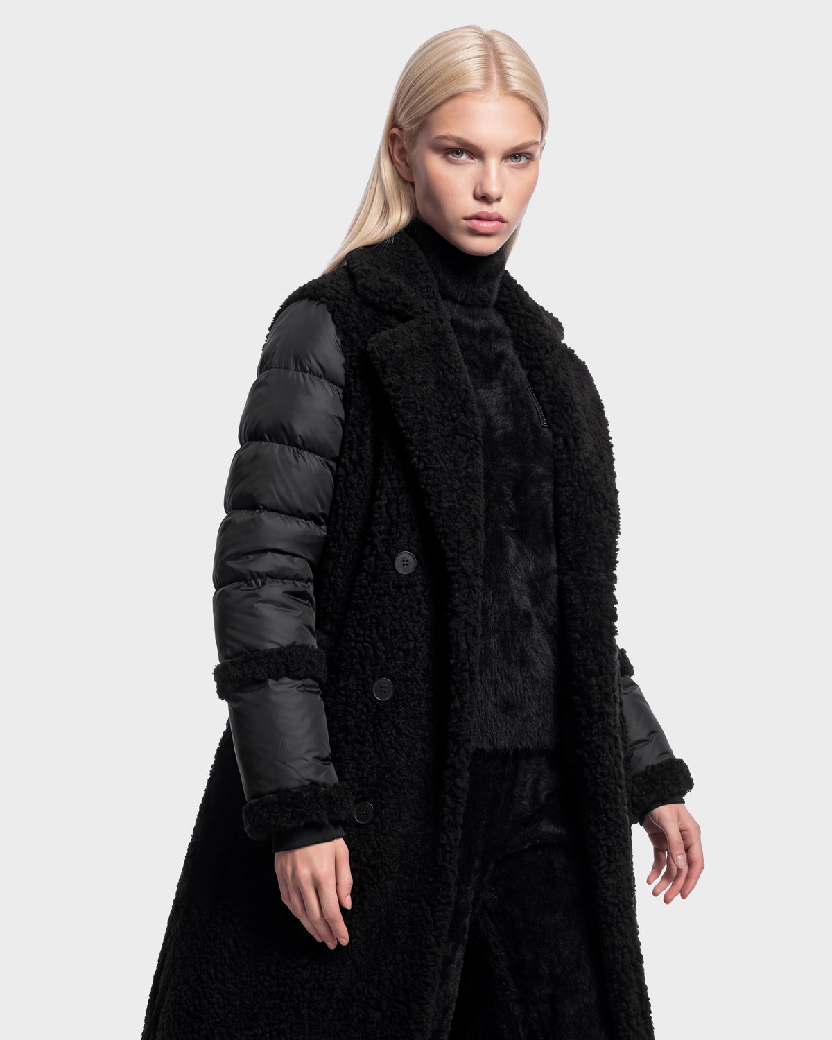 A woman with long blonde hair wears the Rudsak NOELLE - 8126971 Black textured hooded coat with shiny quilted sleeves and black pants, standing against a plain light gray background.