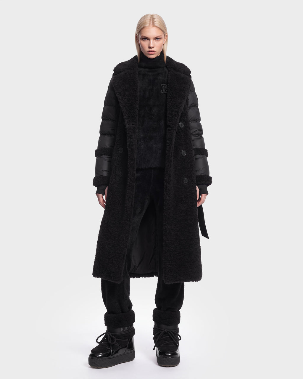 A person with straight blonde hair wears the Rudsak NOELLE - 8126971 Black long faux-fur coat with puffer sleeves, paired with black pants, black boots, and a high-neck top, standing against a plain white background.