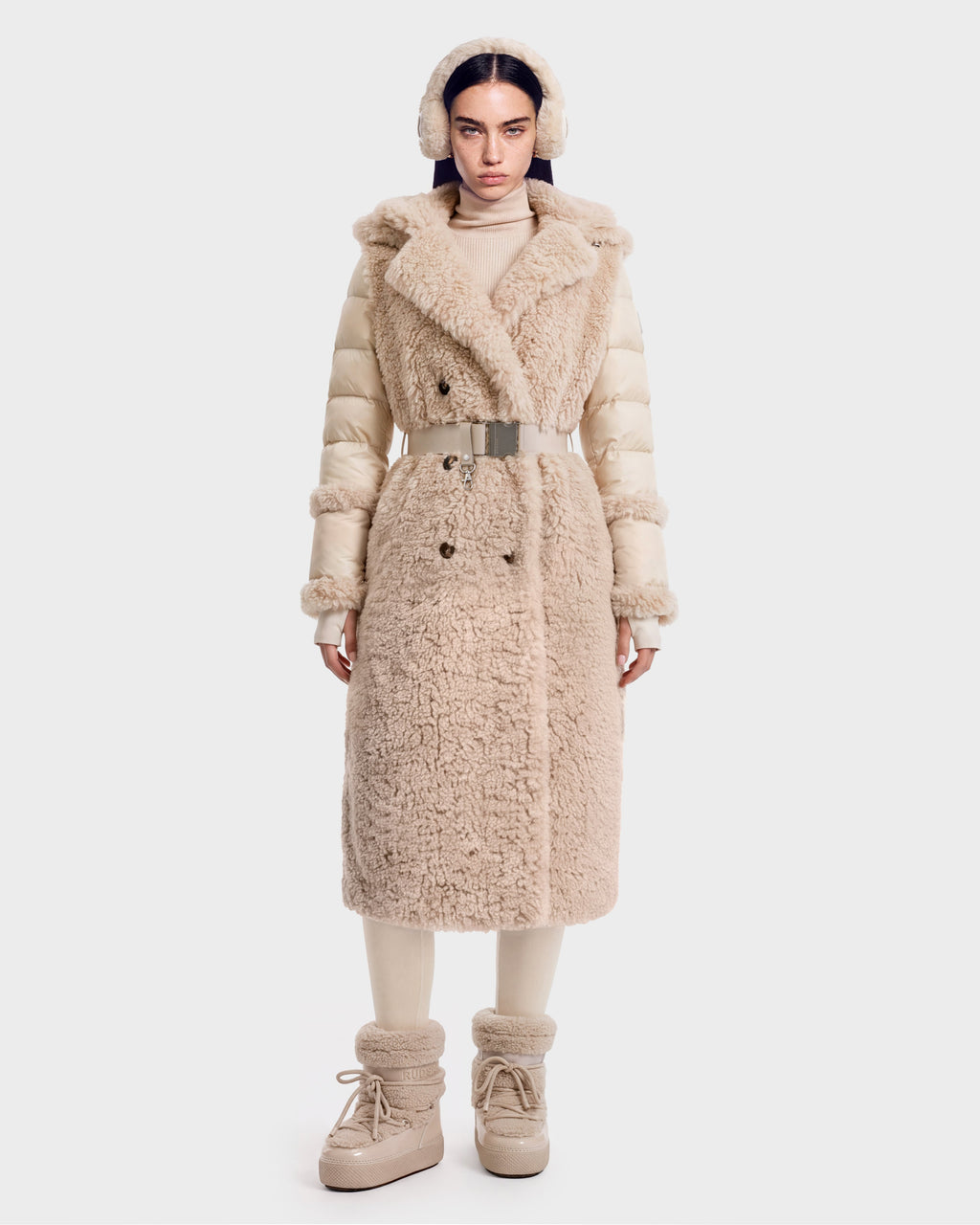 A woman stands facing forward, wearing the Rudsak NOELLE - 8126971 Cream plush faux fur coat with a belt, paired with matching ear muffs, boots, and a turtleneck. The outfit is cozy and winter-appropriate, featuring soft textures and neutral tones.