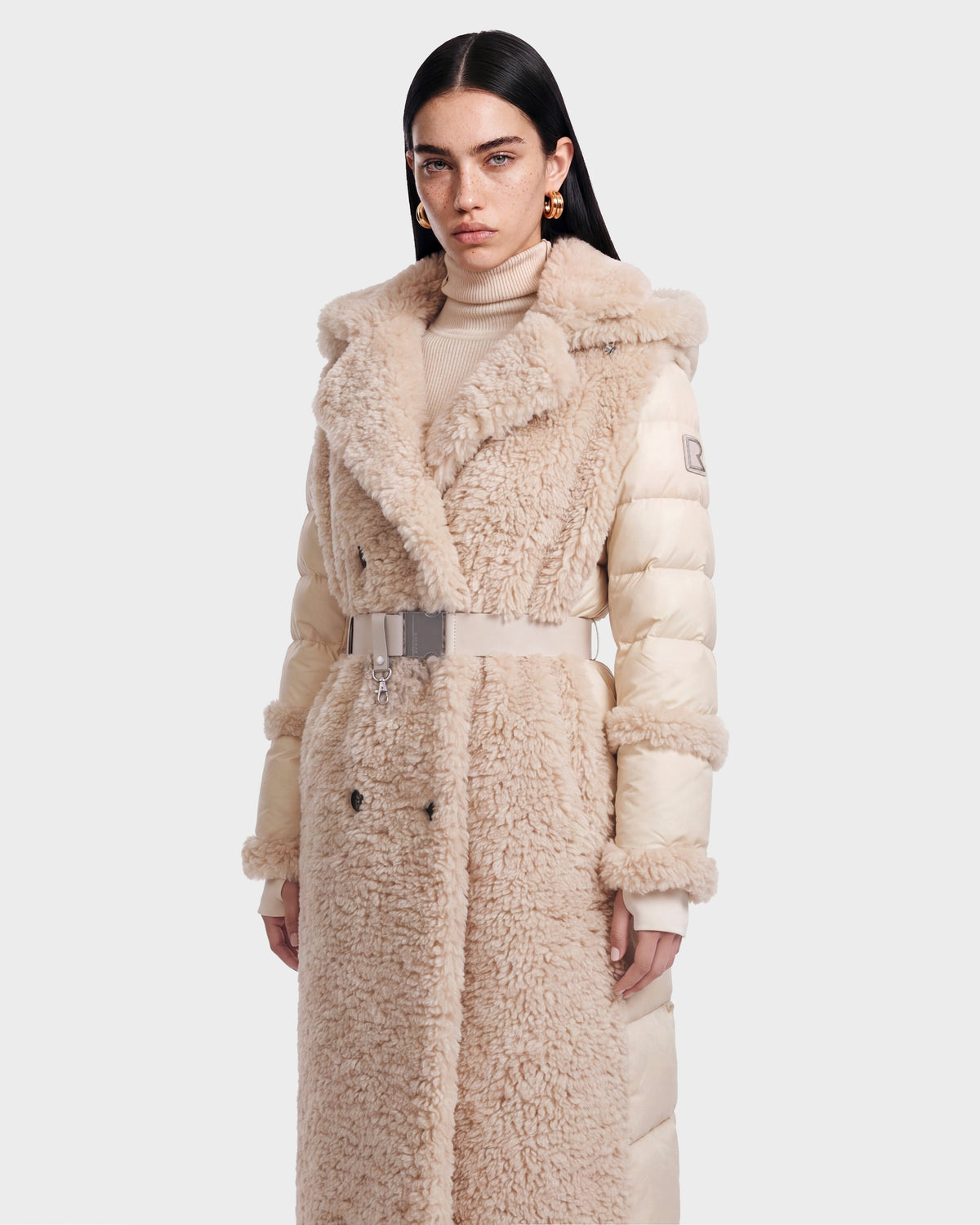 A woman wearing the NOELLE - 8126971 Cream long plush faux fur coat by Rudsak, featuring a belted waist, stands against a plain white background. She has straight dark hair and is accessorized with gold earrings and a beige turtleneck sweater.